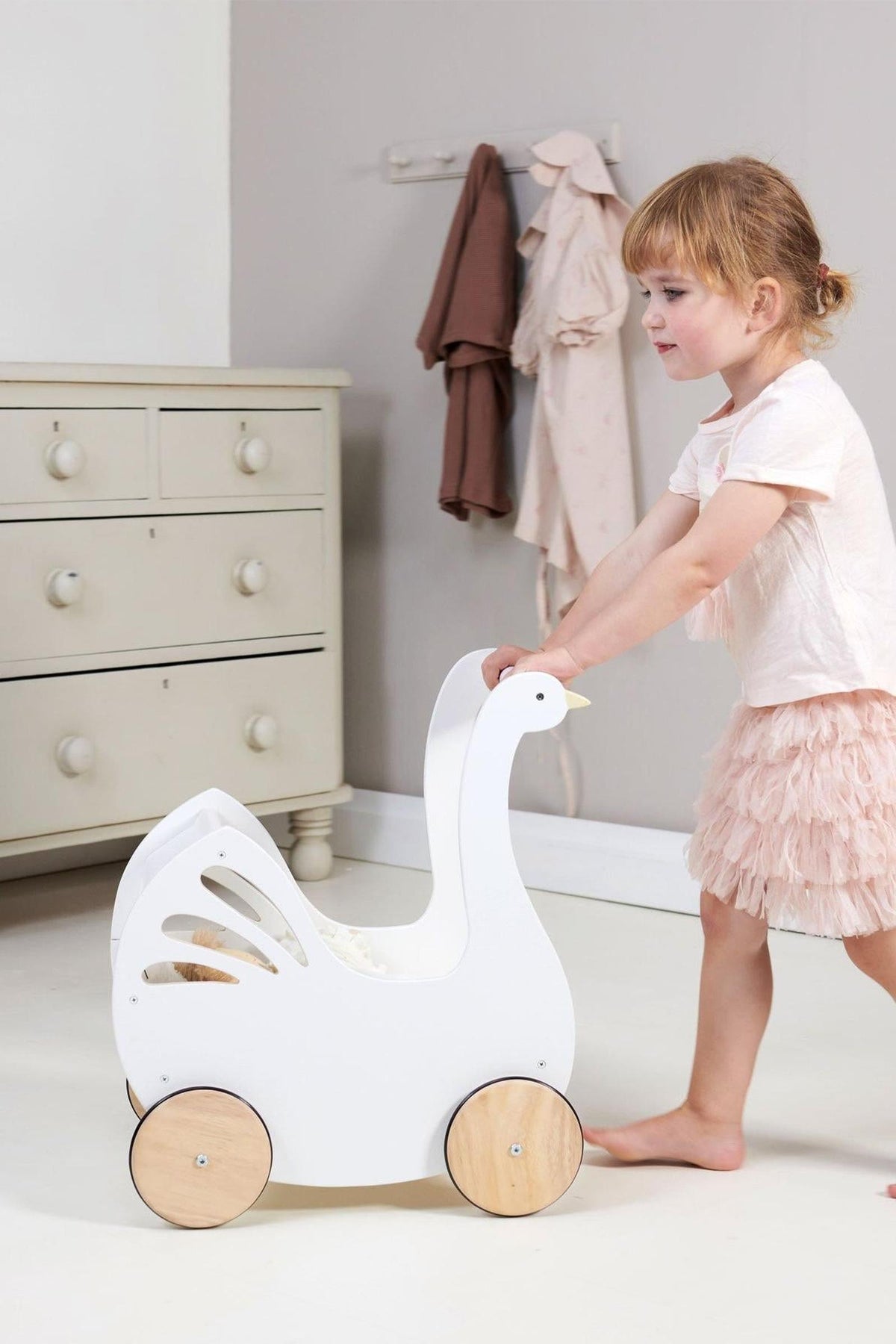 A young child pushes a white wooden baby carriage shaped like a swan, featuring natural wooden wheels. The setting includes a light-colored room with soft textiles in the background.