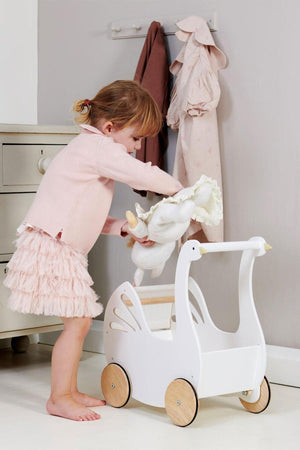A young child in a pink sweater and ruffled skirt is placing a stuffed animal into a white wooden baby carriage shaped like a swan, set against a neutral wall.