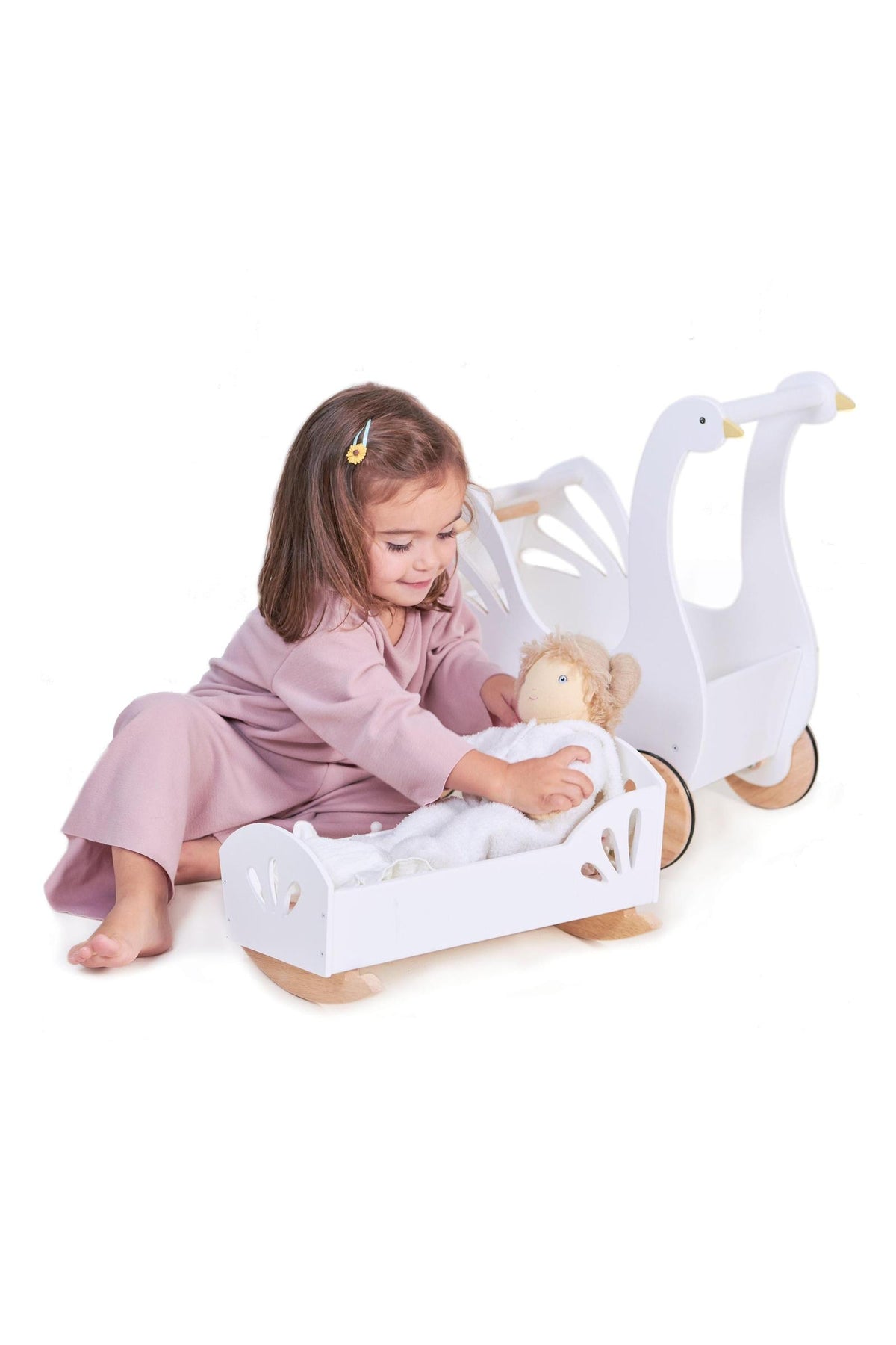A child in pink pajamas plays with a doll in a white swan-themed wooden dolly bed with cutout details.