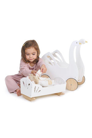 A child plays with a Tender Leaf Sweet Swan Dolly Bed, featuring white wood and natural rockers, alongside a swan-shaped.