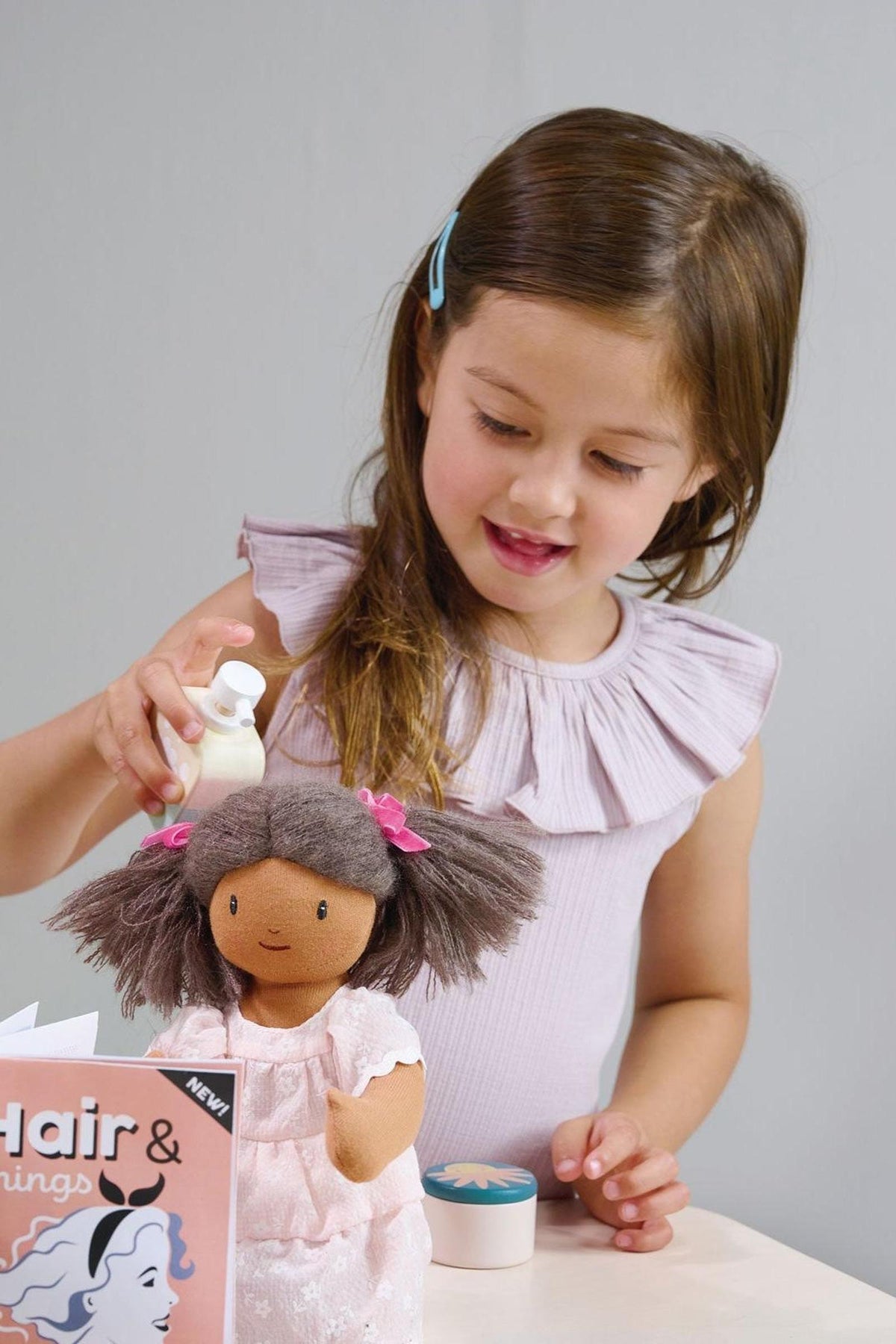 A young girl with long brown hair styles a dolls hair, using a small bottle. The scene features soft colors and a playful atmosphere, showcasing imaginative role play.