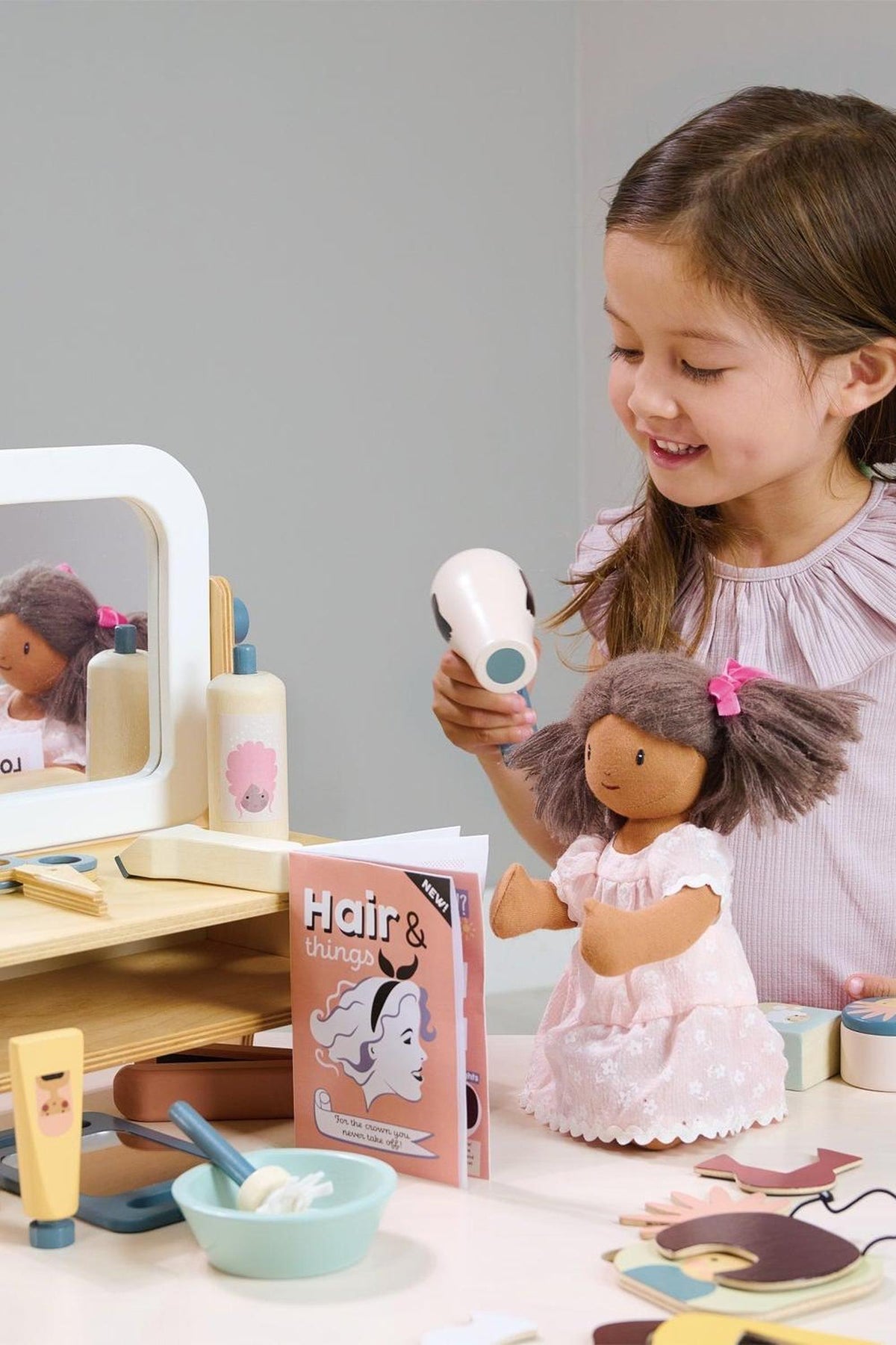 A young girl smiles while using a wooden hairdryer on a doll at a hair salon playset.