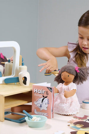 A young girl styles a dolls hair at a wooden hair salon playset, featuring a tabletop mirror and accessories. The soft colors and natural materials promote imaginative play.