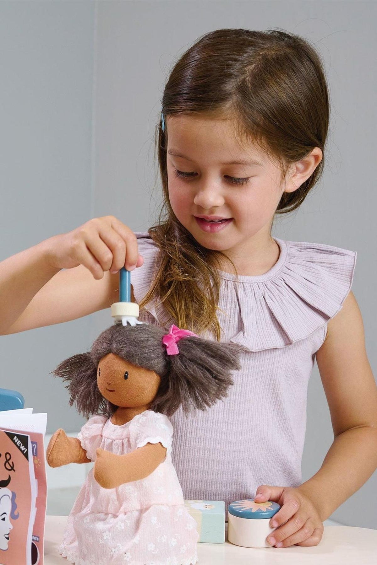 A young girl styles a wooden doll with brown hair and a pink dress, using a blue hairbrush.