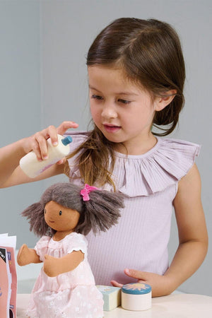A young girl with long brown hair, wearing a light purple top, pretends to style a dolls hair using a spray bottle.