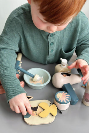 A young child plays with a wooden hair salon toy set, featuring various colorful accessories and a small bowl, all designed for imaginative role play.