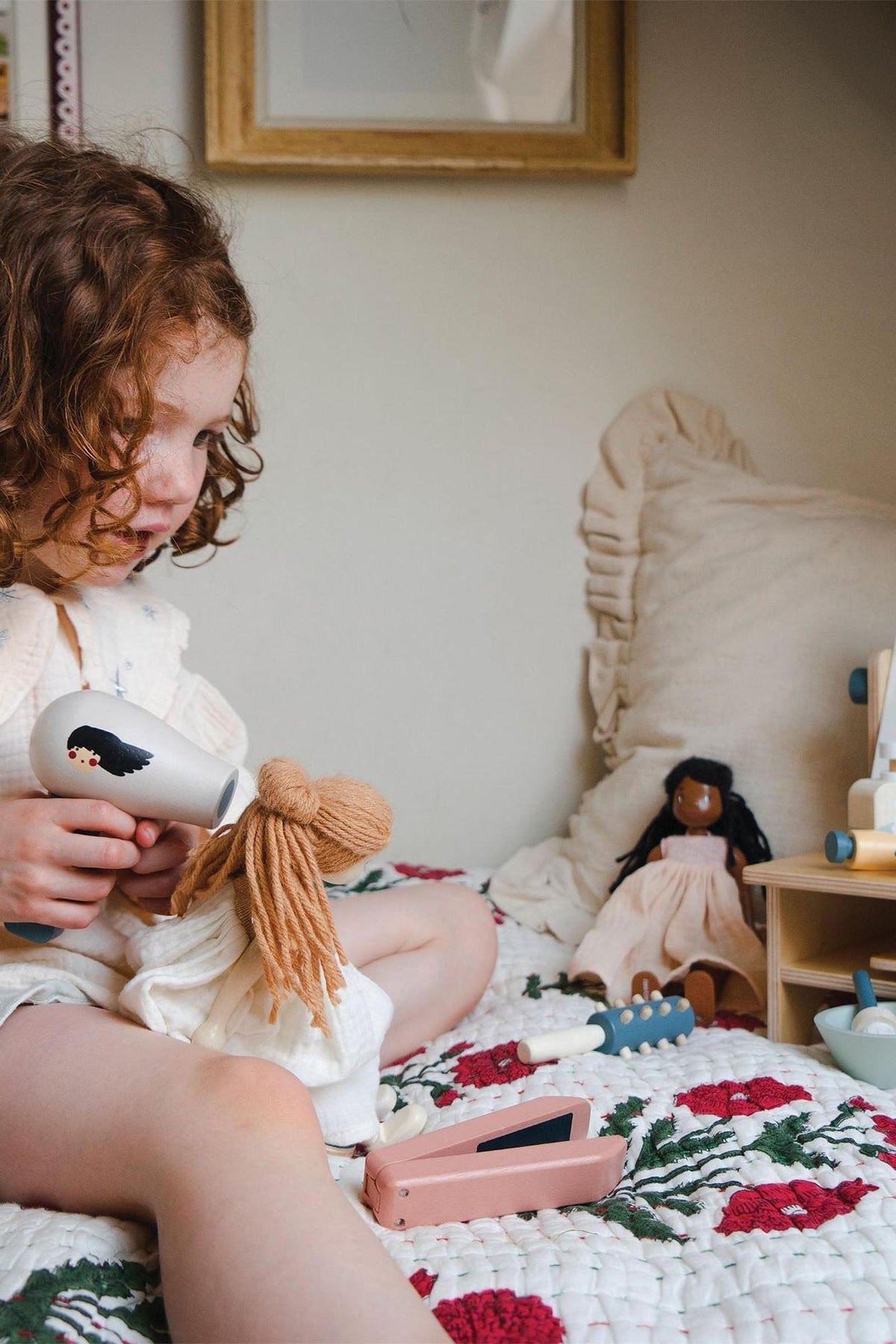 A young child with curly hair plays with a doll on a floral quilt, using a toy hairdryer while another doll sits nearby.