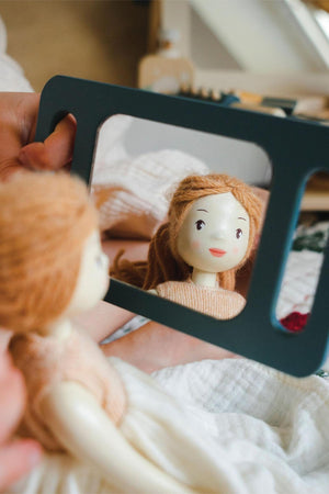 A child holds a wooden doll in front of a tabletop mirror, reflecting the doll’s smiling face. The scene features soft, natural colors and a cozy, playful setting.