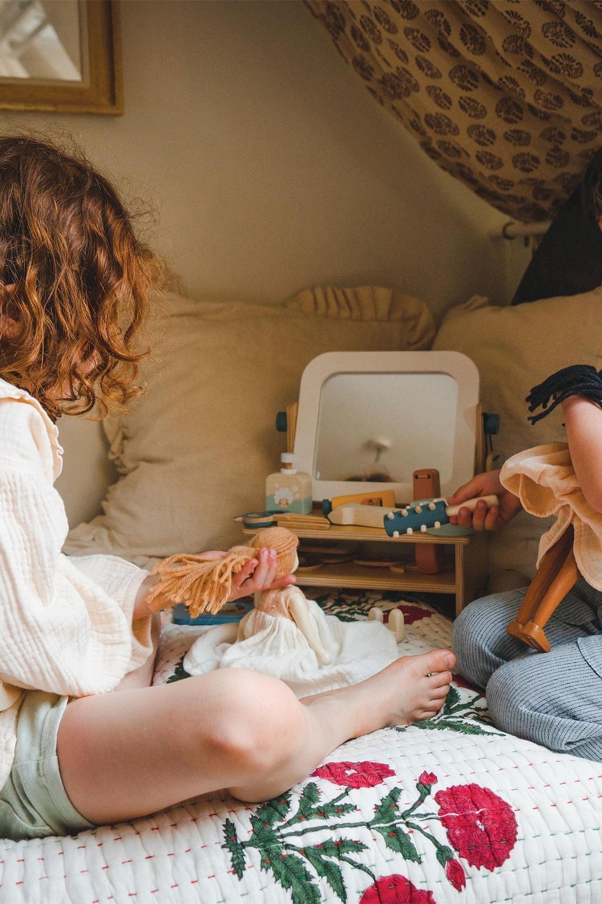 Two children sit on a bed, engaged in imaginative play with a wooden hair salon set featuring a mirror and tools.