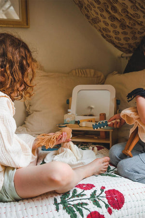 Two children sit on a bed, engaged in imaginative play with a wooden hair salon set featuring a mirror and tools.