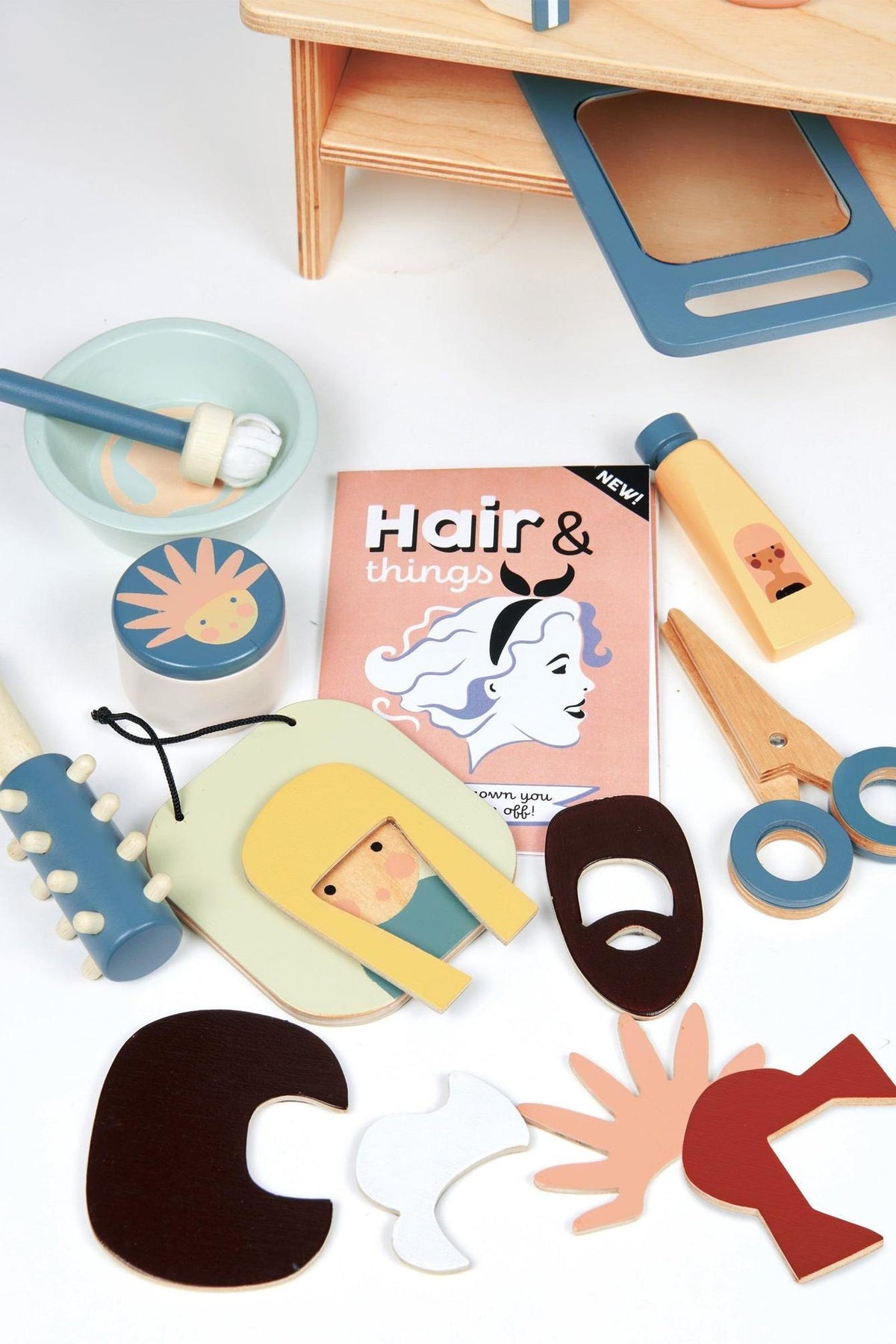 A colorful assortment of wooden hair salon accessories, including scissors, hair pieces, and a playful styling guide, is arranged on a light wooden surface, ideal for imaginative play.