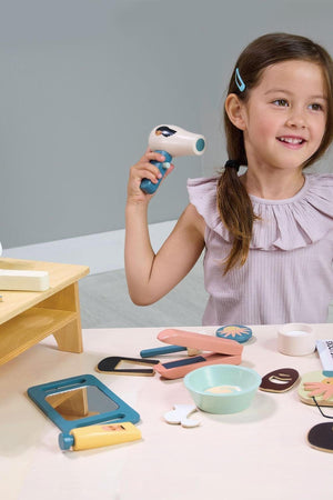 A young girl smiles while holding a toy hairdryer, surrounded by colorful wooden hair salon accessories.