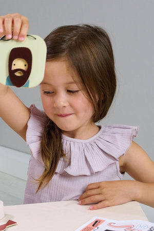 A young girl with long brown hair holds a tabletop mirror with a playful design, smiling as she engages in imaginative play.