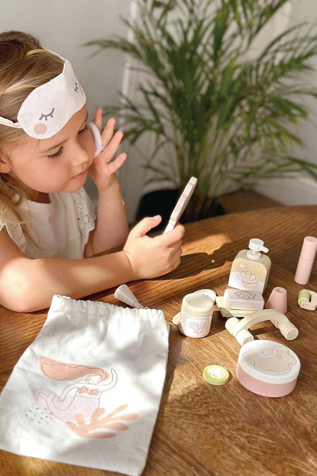 A child wearing a fabric eye mask sits at a wooden table, surrounded by spa-themed toys and a drawstring bag.