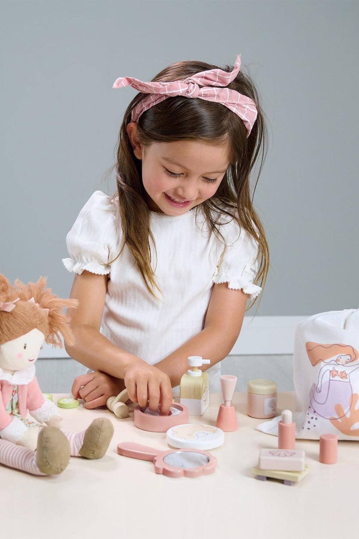 A young girl with a pink headband enjoys playing with a spa retreat toy set, including a mirror and lotion.