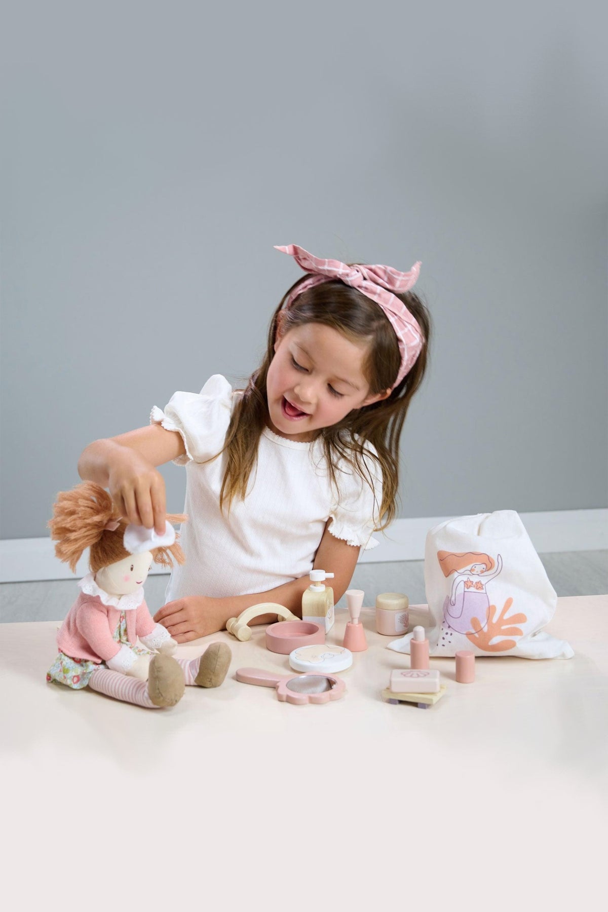 A young girl with a pink bow plays with a plush doll and spa accessories on a light-colored table.