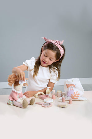 A young girl with a pink bow plays with a plush doll and spa accessories on a light-colored table.