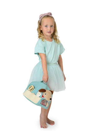 A young girl with curly hair, wearing a light blue top and a tulle skirt, holds a soft, blue toy vet case featuring animal designs.