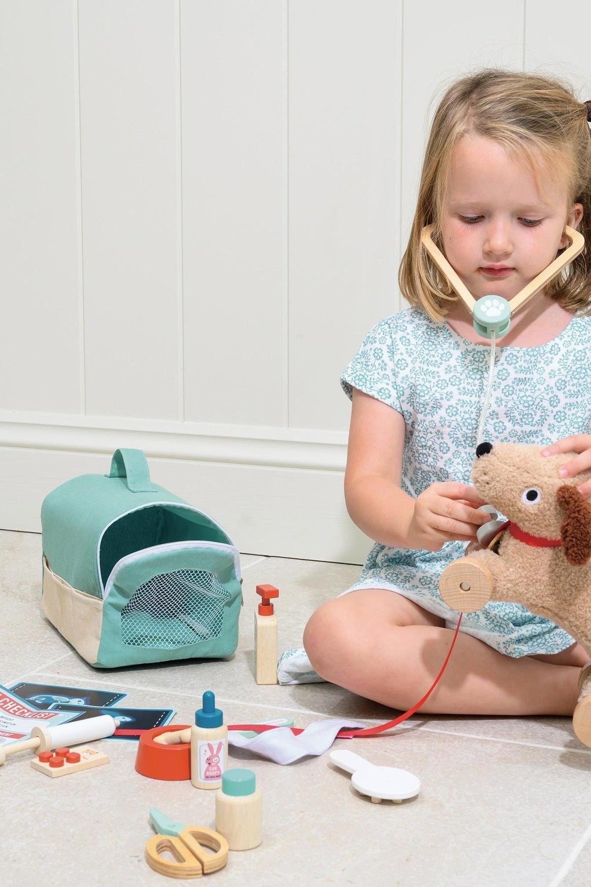 A young girl uses a wooden stethoscope on a plush dog while seated on the floor, surrounded by veterinary tools and a soft carry case in a light, bright setting.