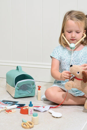 A young girl uses a wooden stethoscope on a plush dog while seated on the floor, surrounded by veterinary tools and a soft carry case in a light, bright setting.