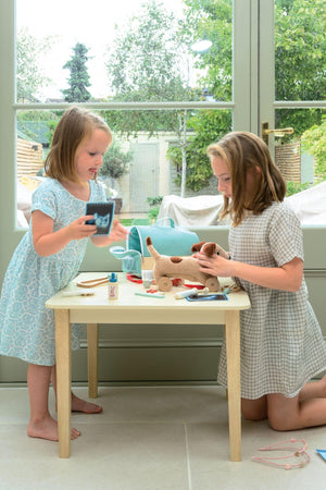 Two children play at a light-colored table, one holding a toy while the other examines a wooden animal. The setting features large windows with greenery outside, creating a bright atmosphere.