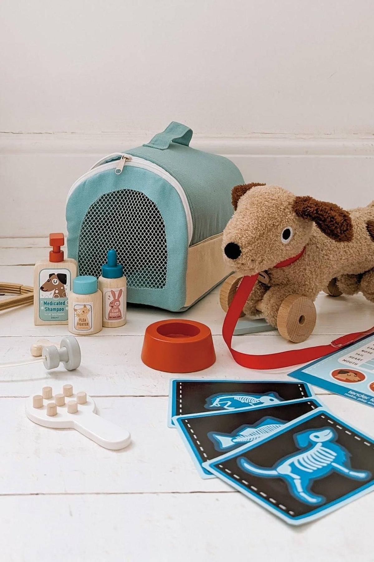 A soft turquoise pet carrier sits alongside a plush brown dog toy, wooden veterinary tools, and pet care cards. The eco-friendly materials enhance imaginative play for young animal lovers.