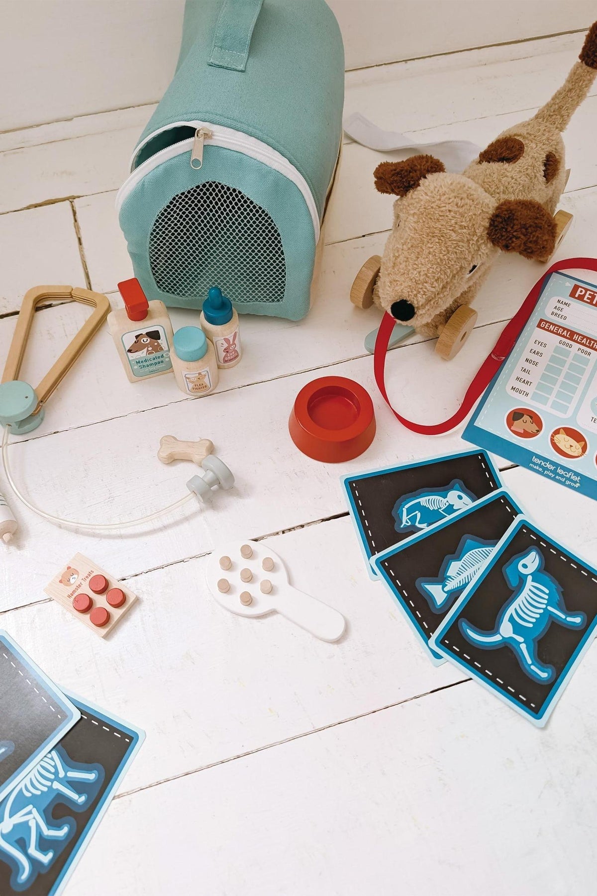 A soft aqua vet case sits beside a plush dog, with wooden veterinary tools, pet care cards, and colorful accessories scattered on a light wooden surface, inviting imaginative play.