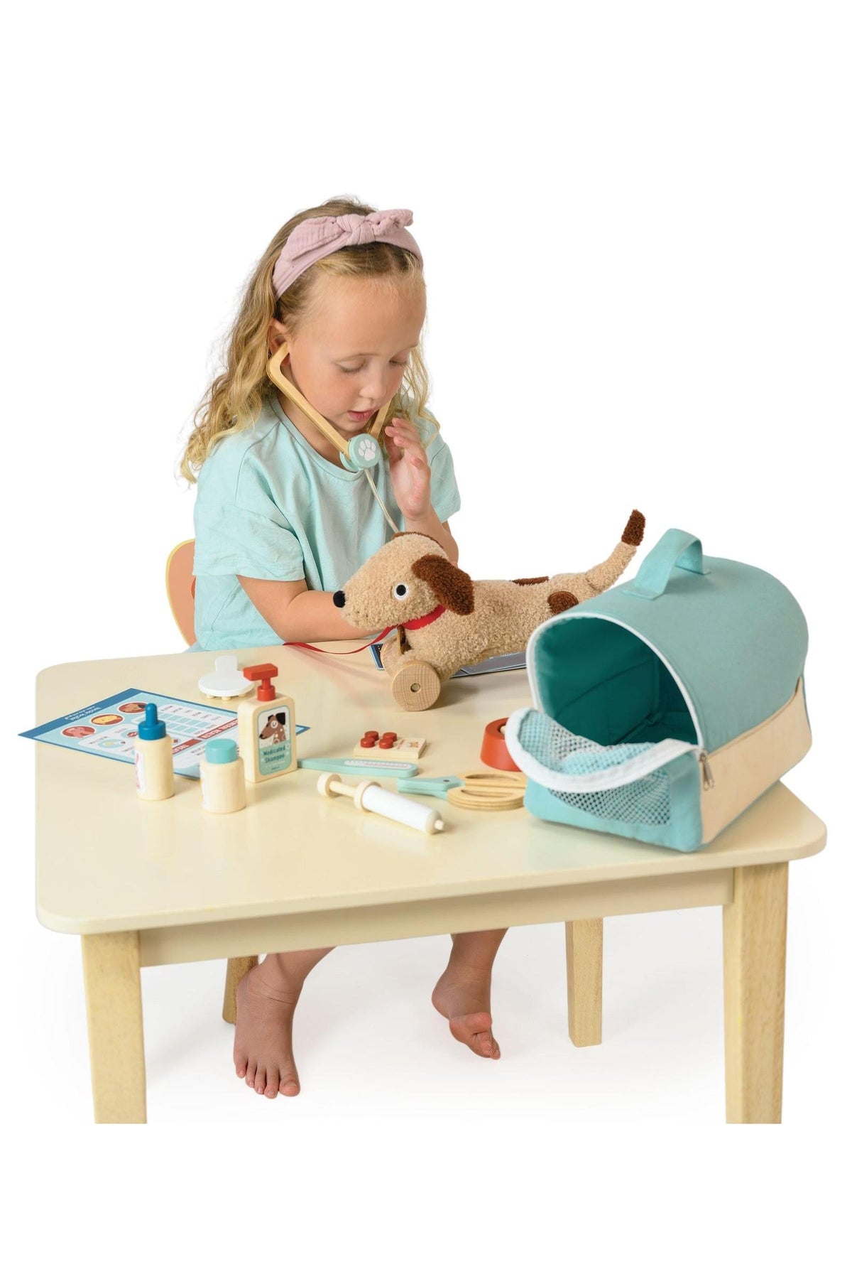 A child with curly hair wears a light blue shirt and headband, examining a plush dog on a table.