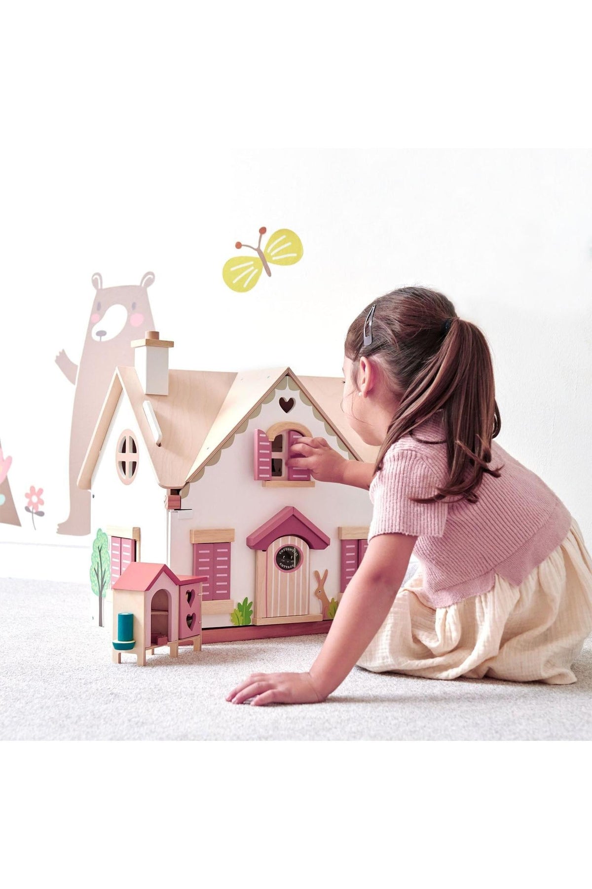 A child interacts with a colorful wooden dollhouse featuring pink shutters and a natural wood roof, set against playful wall.