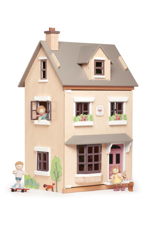 A wooden dollhouse with a light plywood façade and gray roof features colorful window boxes and two figurines playing outside.