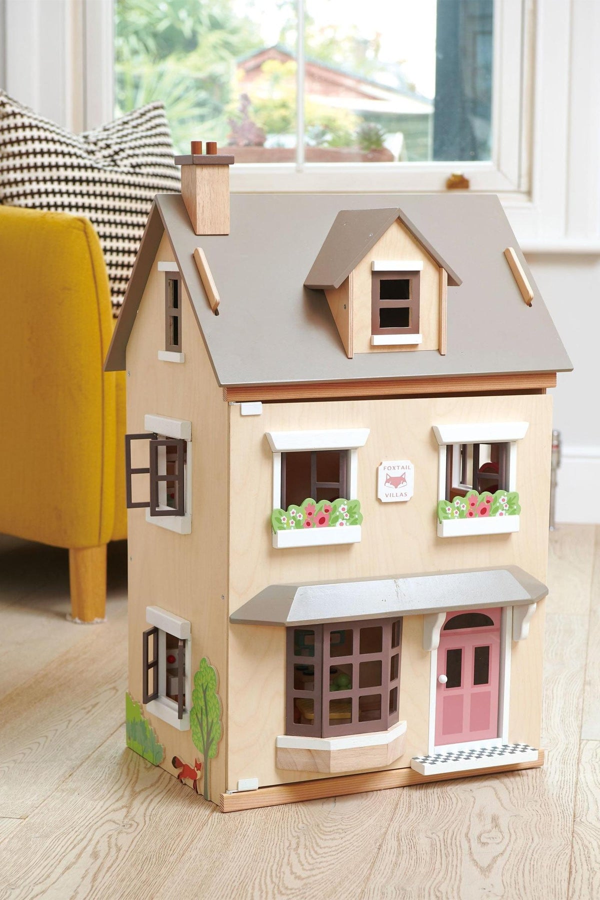 A wooden dollhouse with a beige façade, gray roof, and colorful flower boxes, set in a bright living space.