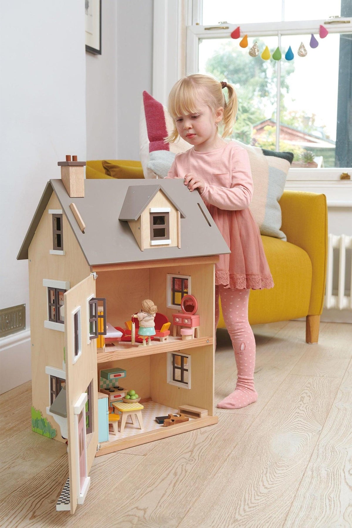 A young girl interacts with a wooden dollhouse, featuring a neutral facade and colorful interior accessories, in a bright living.