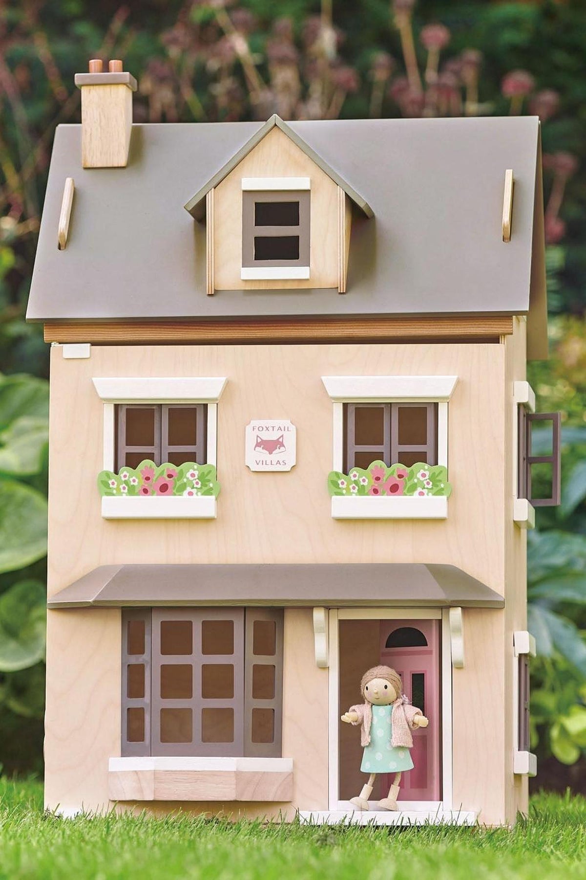 A wooden dollhouse with a natural plywood finish, featuring pastel colors, flower decorations, and a miniature figure at the door.