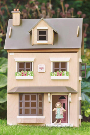 A wooden dollhouse with a natural plywood finish, featuring pastel colors, flower decorations, and a miniature figure at the door.