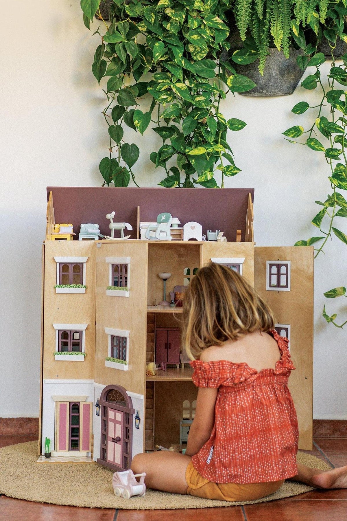 A young child plays with a wooden dollhouse featuring multiple rooms and detailed furniture. Lush green plants adorn the background, enhancing the inviting atmosphere of imaginative play.