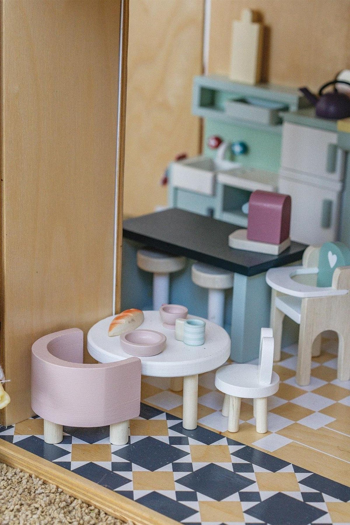 A detailed view of a miniature wooden kitchen scene featuring a round table, pastel-colored chairs, and various kitchen accessories. The setup reflects a charming and playful design suitable for children.