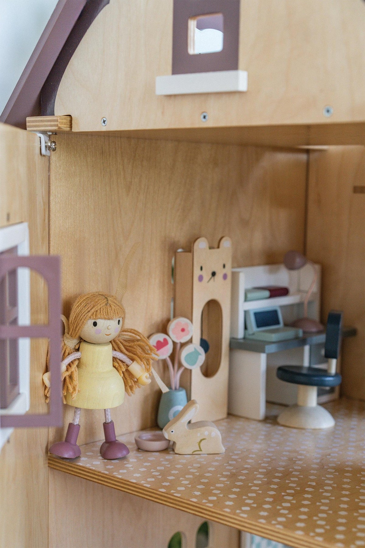 A wooden dollhouse interior features a cheerful girl figurine with braided hair, a desk, and a cute bunny alongside decorative flowers.