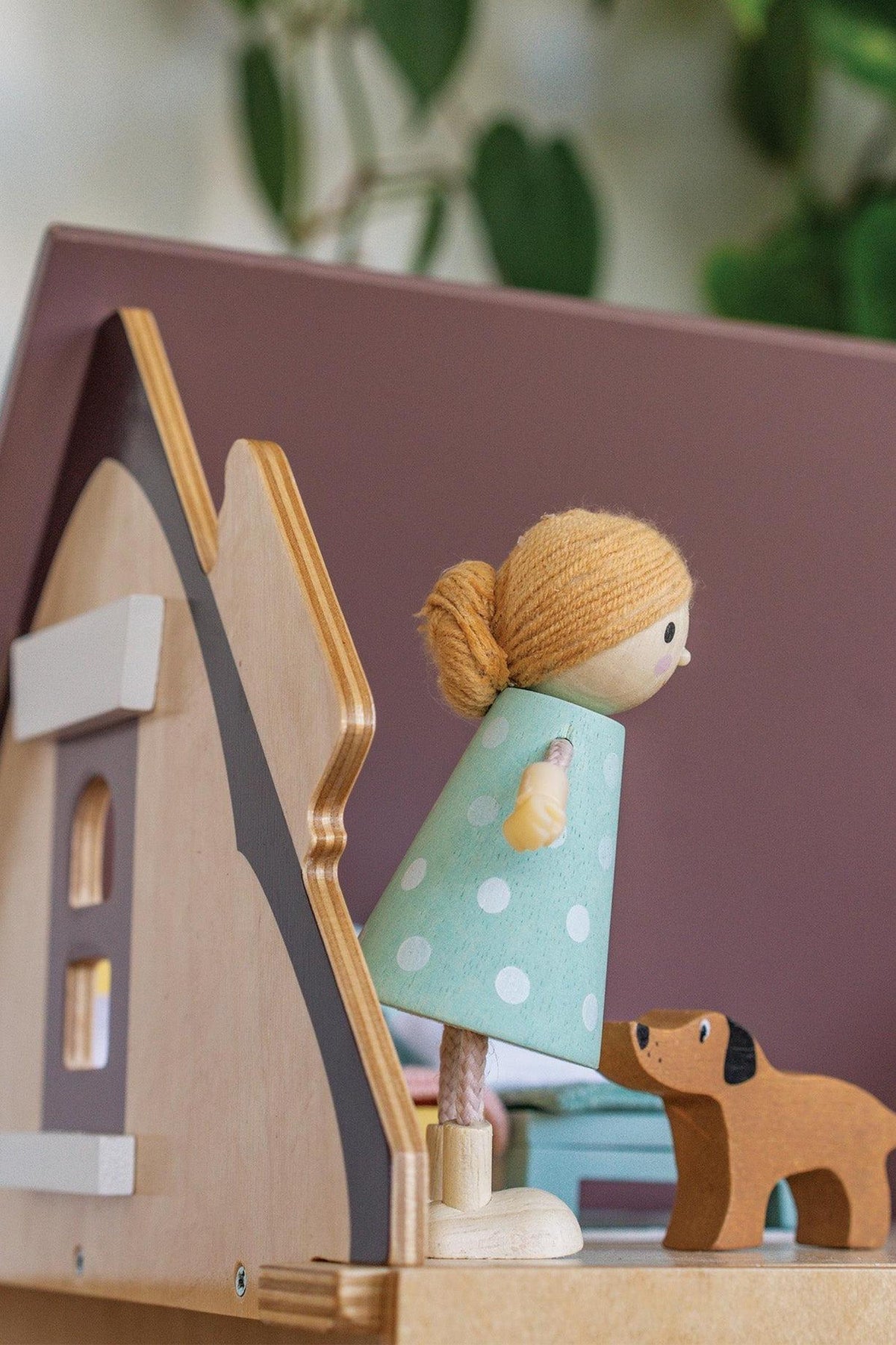 A wooden dollhouse scene features a charming figure in a polka dot dress and a small brown dog, showcasing sustainable materials and imaginative play. Greenery softly decorates the background.