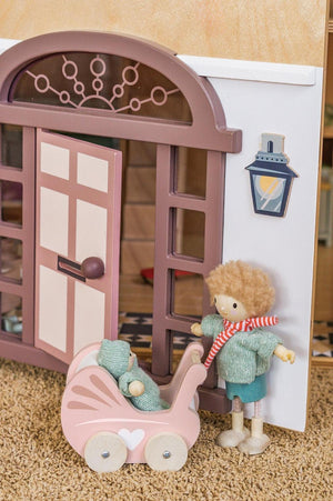 A wooden dollhouse entrance features a pink stroller and a character with curly hair, wearing a green sweater and blue shorts.