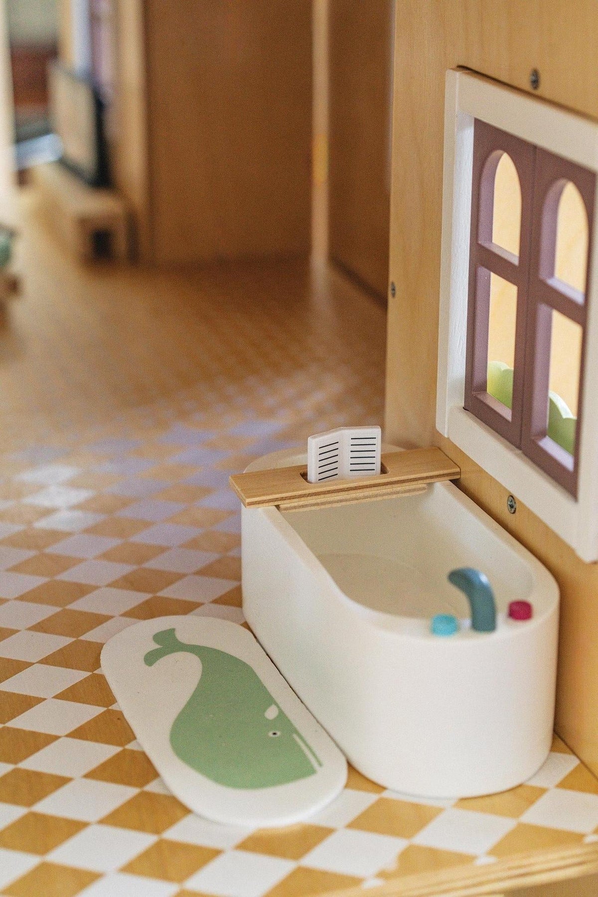 A white wooden bathtub sits on a checkered floor inside a dollhouse, accompanied by a whale-shaped bath mat. The scene features pastel-colored accents and a cozy, inviting atmosphere.