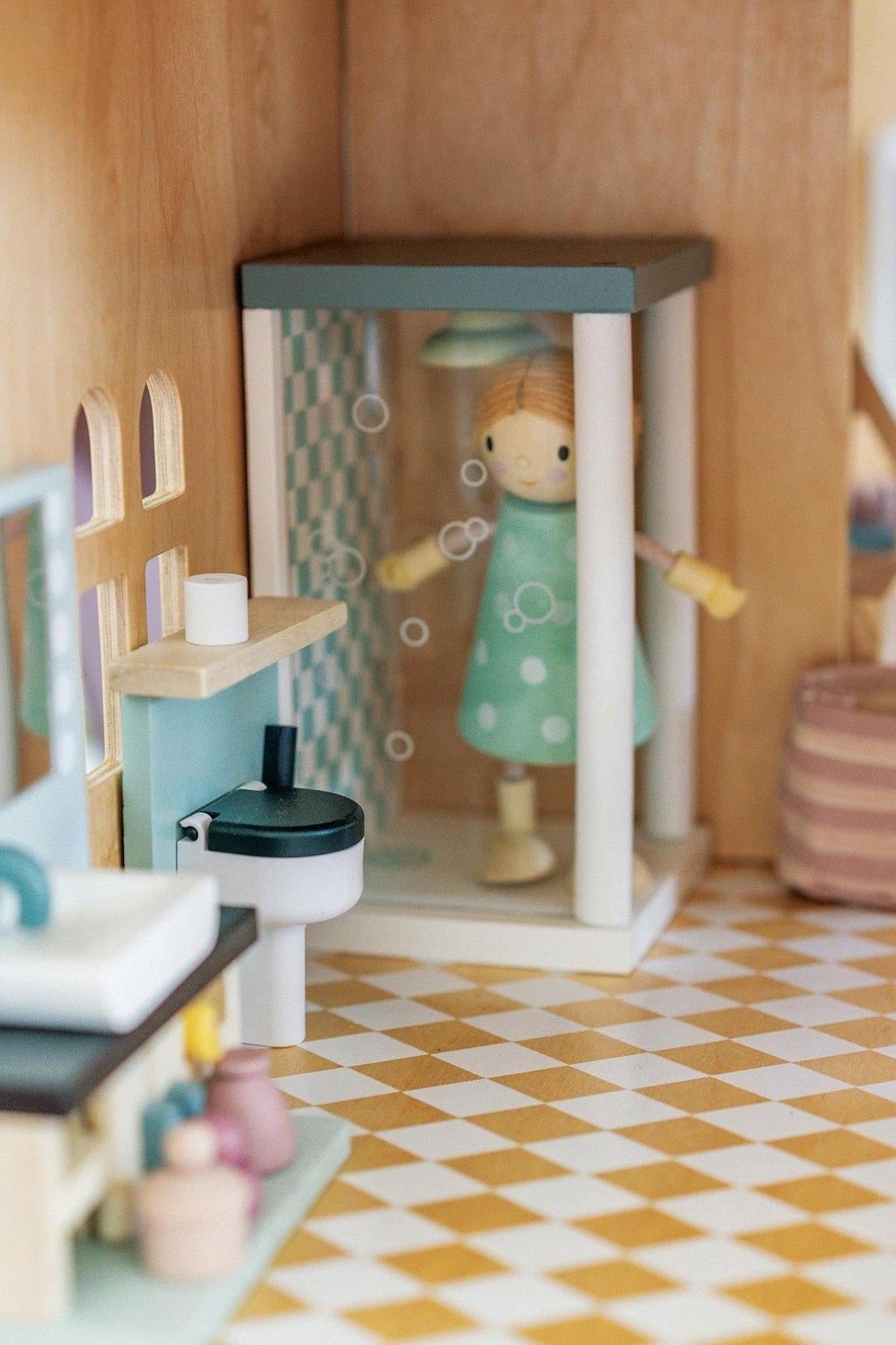 A charming bathroom scene inside a wooden dollhouse features a shower with bubbles, a colorful doll in a polka-dotted dress, and a stylish sink.