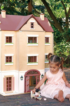 A child in a pink dress plays with a toy dog in front of the Tender Leaf Wooden Toys Fantail Hall dollhouse, showcasing its elegant wooden design and charming details.