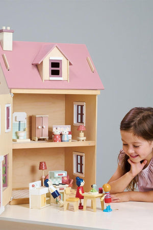 A young girl interacts with a colorful wooden dollhouse featuring a pink roof, miniature furniture, and playful wooden figures.