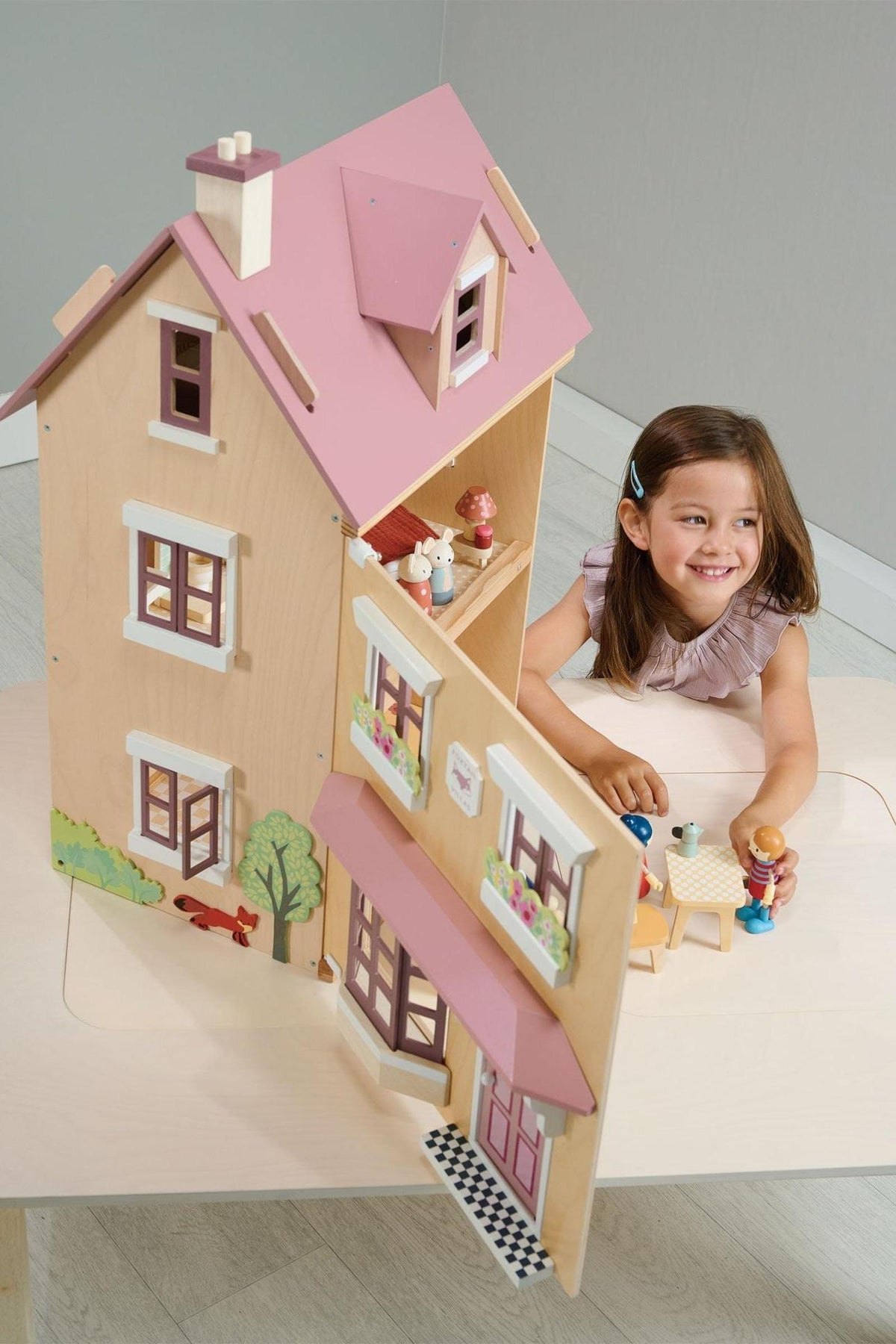 A young girl plays with a wooden dollhouse featuring a pink roof and colorful figures, set on a light table.