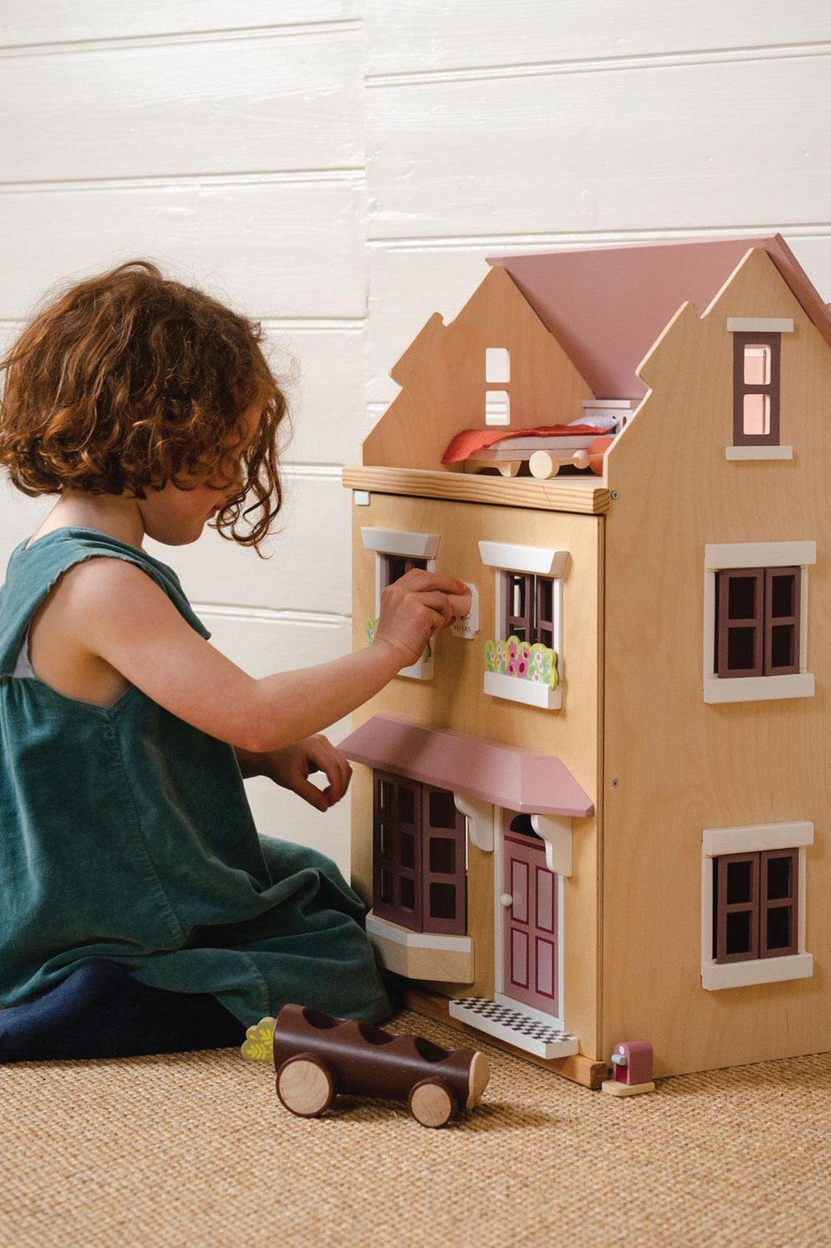 A child interacts with a wooden dollhouse, featuring pastel-colored windows and a pink roof, on a natural fiber mat.