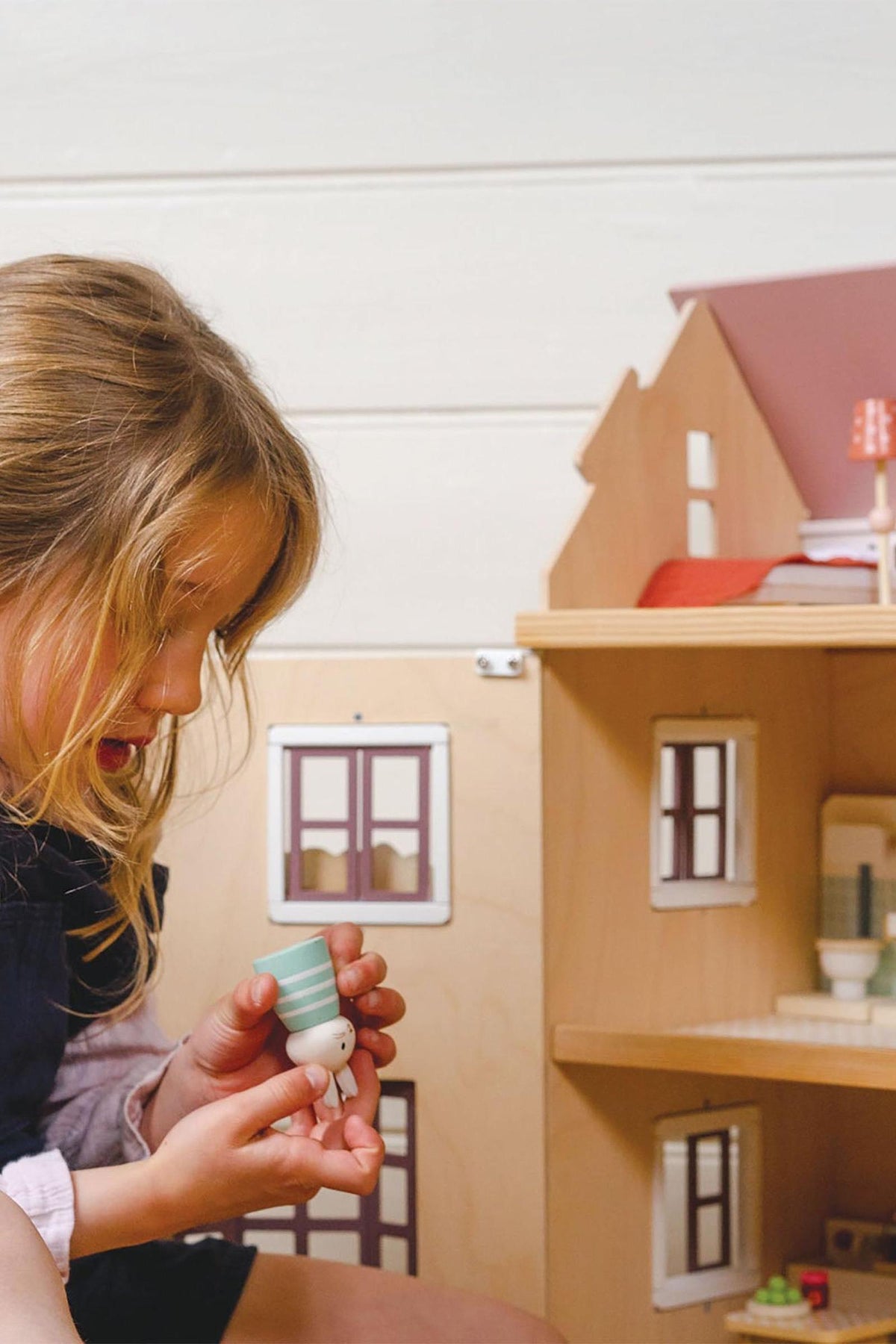 A child holds a small wooden figure in front of a dollhouse, showcasing its intricate design and colorful details.