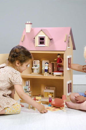 Two children play with a wooden dollhouse featuring a pink roof, colorful furniture, and miniature characters.