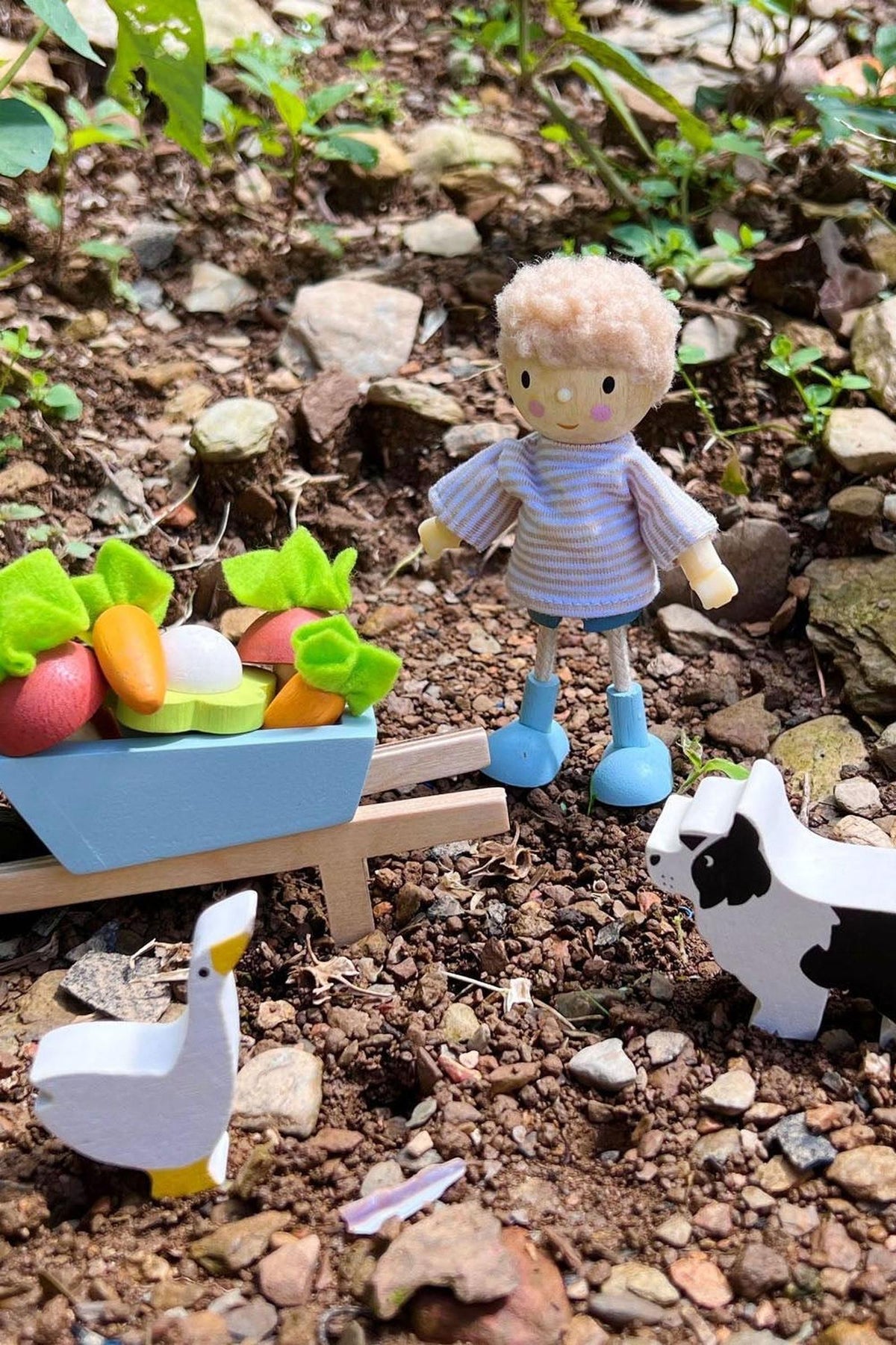 A small wooden figure with curly hair stands beside a colorful cart filled with toy vegetables, surrounded by a natural setting with rocks and greenery, creating a playful garden scene.