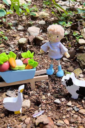 A small wooden figure with curly hair stands beside a colorful cart filled with toy vegetables, surrounded by a natural setting with rocks and greenery, creating a playful garden scene.