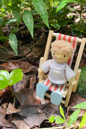 A wooden doll with a fluffy hairstyle sits in a miniature deck chair among green leaves and brown foliage, creating a vibrant outdoor play scene.